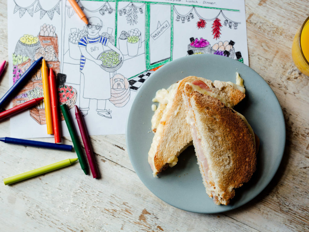 bird’s eye view image of a ham and cheese toasted sandwich on a grey plate and a glass of orange juice on top of a children’s colouring in sheet and crayons in various colours such as green, purple, blue, orange and red on top of a wooden table