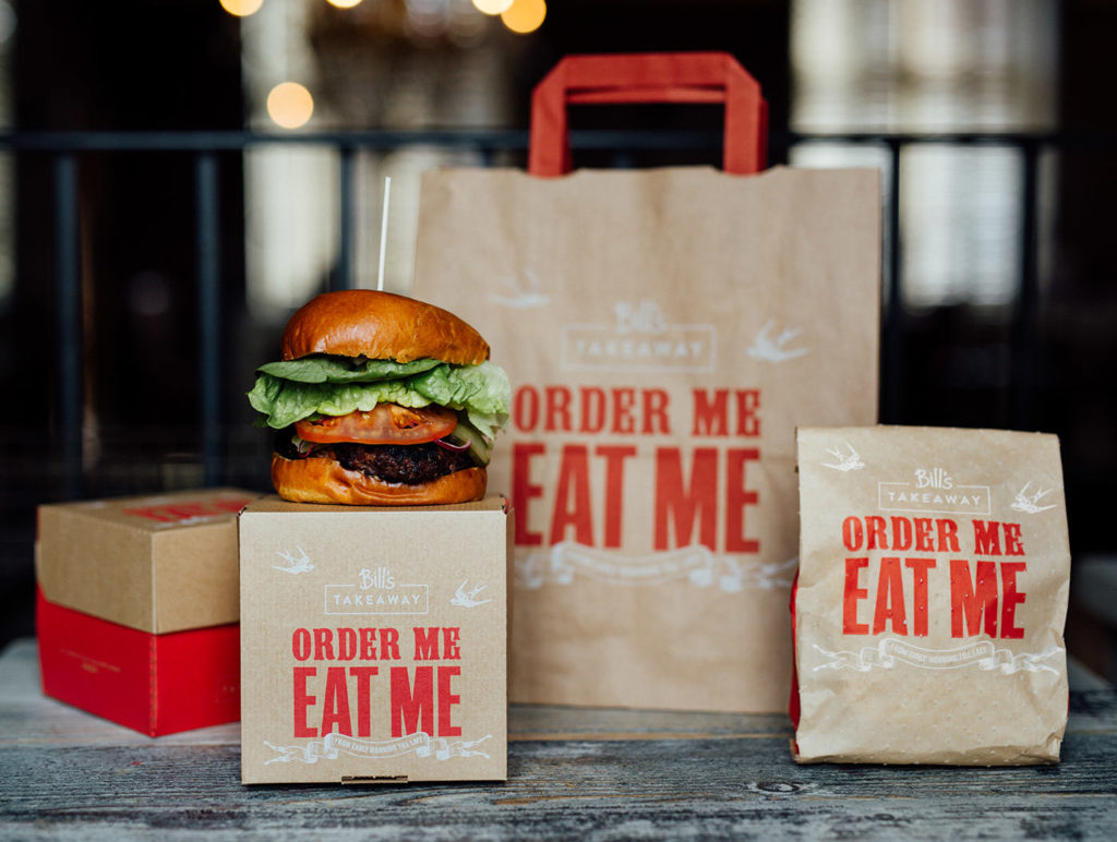 A burger consisting of bread, meat, lettuce and tomato sitting on top of a branded burger box with the Bill’s logo in white and red text underneath it reading ‘order me, eat me’ surrounding by other branded packaging including a small bag