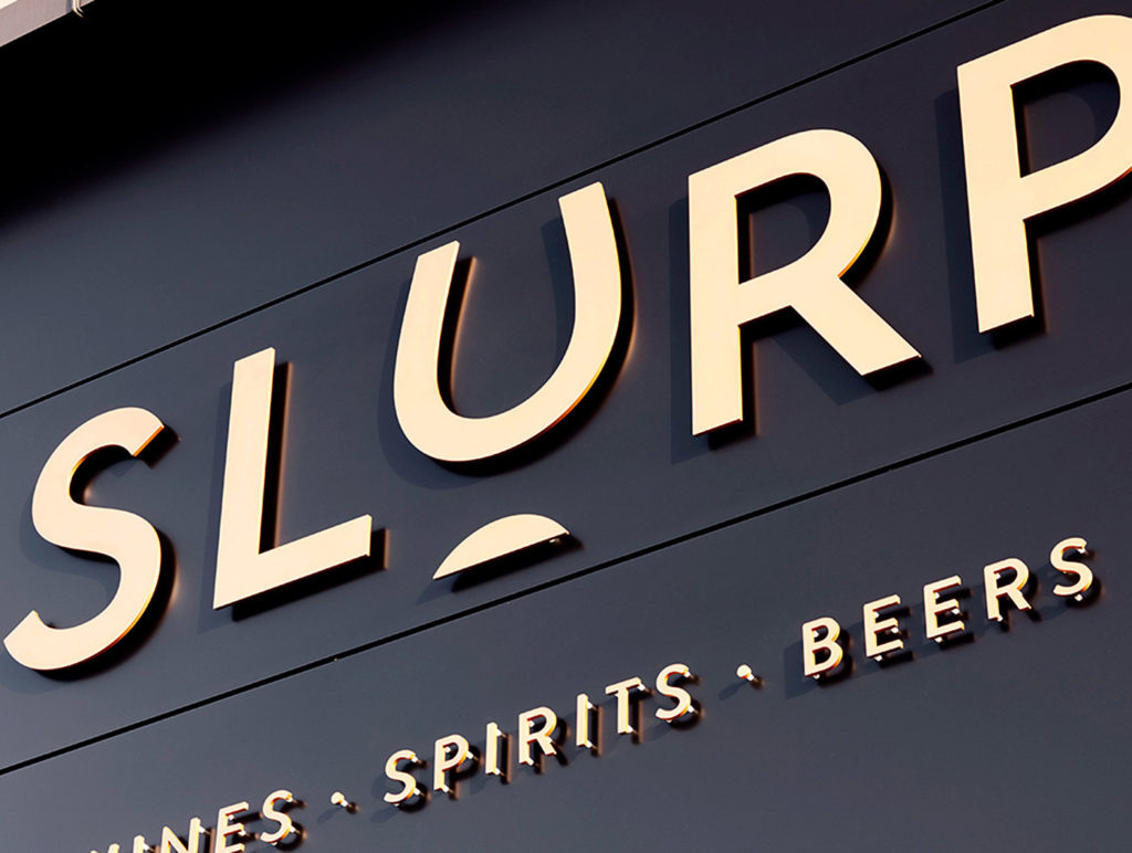 close-up photograph of the ‘Slurp’ logo in bright gold letters and the words ‘Wine, Spirits, Beers’ underneath on the outside of a shop against a black background