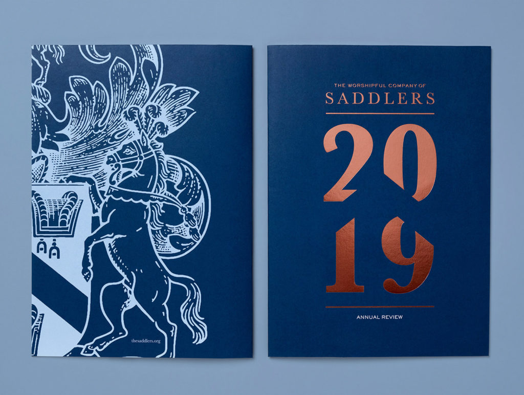 back of the brochure showing a section of the coat of arms in blue against a dark blue background on the left, the front of the brochure with foil words 'The Worshipful Company of Saddlers' and the numbers '2019' on the front and the words 'annual report' in white on the right