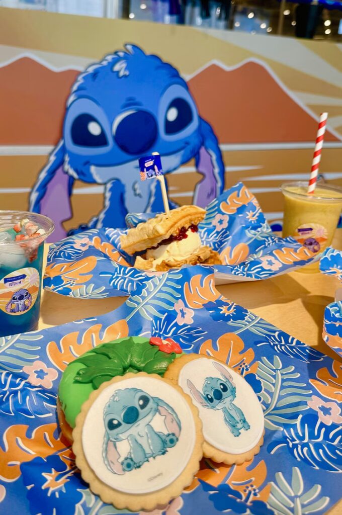An illustrated promotional image for “Stitch Café,” themed around Disney’s Stitch character. The design features a pastel colour palette with a café motif. Stitch is illustrated in a playful pose, surrounded by desserts, tea sets, and café signage. The setting has a soft, inviting feel with pink, blue, and cream tones. Decorative elements such as cupcakes, biscuits, and floral patterns enhance the charming, whimsical café atmosphere.