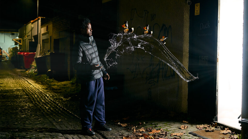 Side profile of a man standing in a dark alleyway who is looking surprised as a row of floating duck approaches him