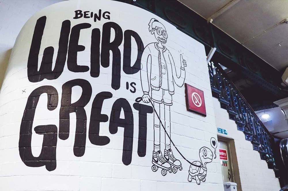 Photo of a wall art with the text 'Being weird is great'. Beside the text is a doodle of a standing girl holding a turtle on a leash