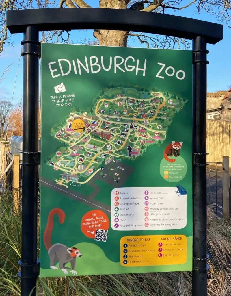 Illustrated signboard at Edinburgh Zoo showing a colourful map of the zoo layout with paths, animal enclosures, and key visitor facilities. The map is accompanied by a clear legend indicating locations for toilets, cafés, picnic areas, and first aid, as well as icons for steep paths and accessible routes. A “You Are Here” marker and playful animal illustrations add to the family-friendly, easy-to-navigate design.